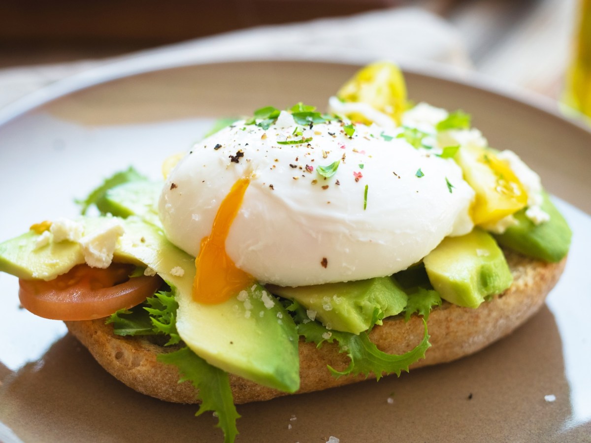 Ein Teller mit einem halben Brötchen, auf dem Avocado und ein pochiertes Ei aus der Heißluftfritteuse liegen. Nahaufnahme.