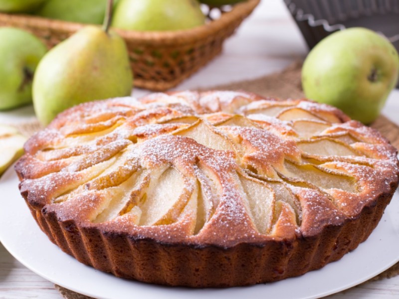 Ein Birnenkuchen mit Puderzucker auf einem Teller. Im Hintergrund liegen frische Birnen.