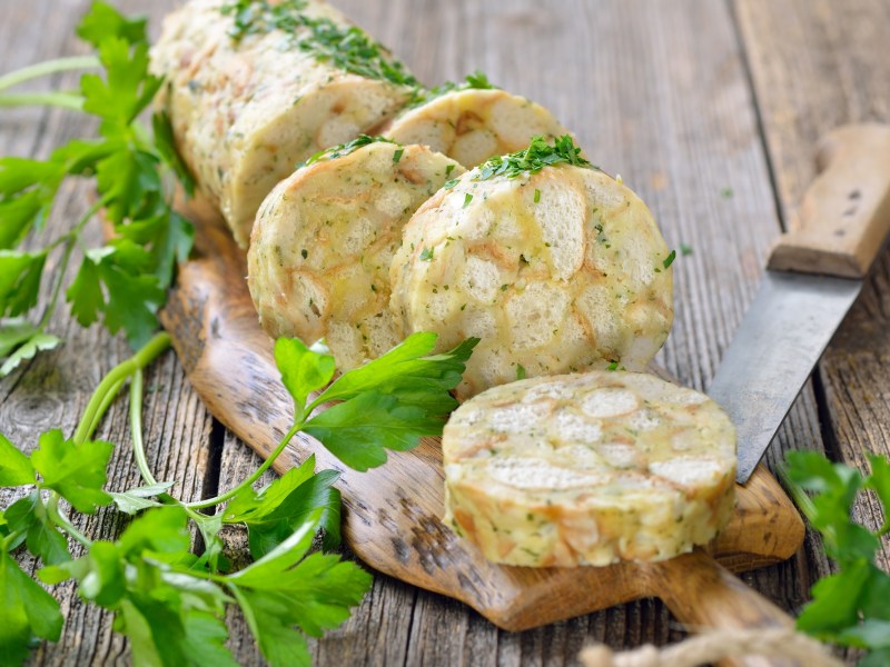 Ein angeschnittener ServiettenknÃ¶del auf einem Brett, daneben ein Messer und Petersilie.