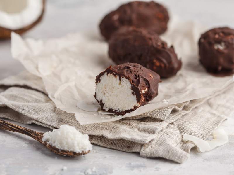 vegane Bounty mit Schokoglasur auf einem StÃ¼ck Papier, daneben eine halbe Kokosnuss und ein LÃ¶ffel Kokosraspeln.