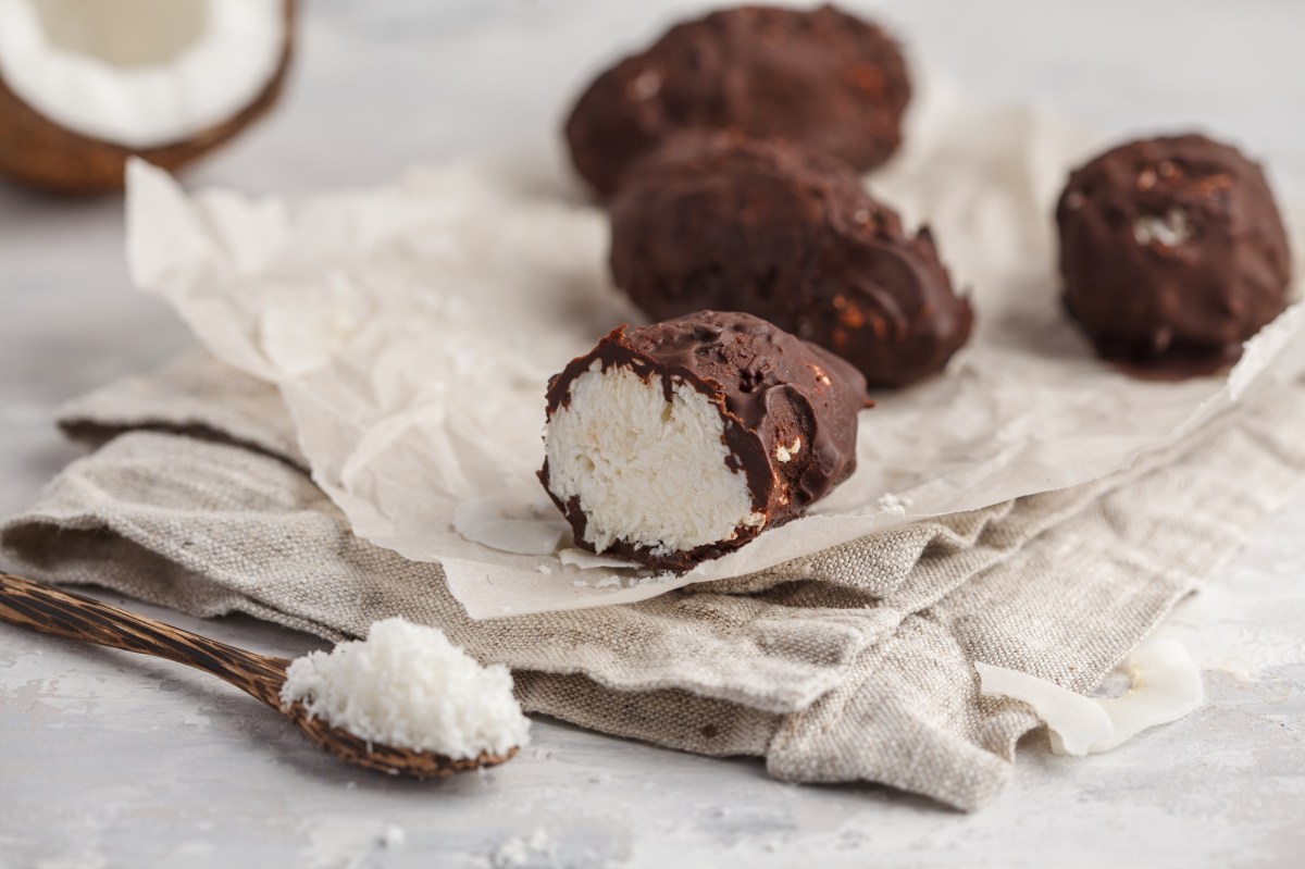 vegane Bounty mit Schokoglasur auf einem StÃ¼ck Papier, daneben eine halbe Kokosnuss und ein LÃ¶ffel Kokosraspeln.