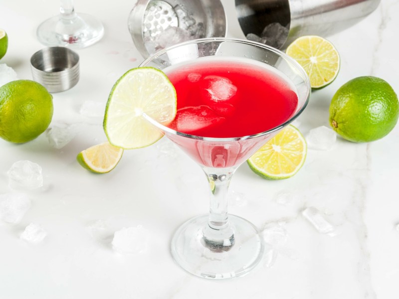 Glas alkohilfreier Cosmopolitan auf einem Tisch mit Limetten und Cocktailshaker