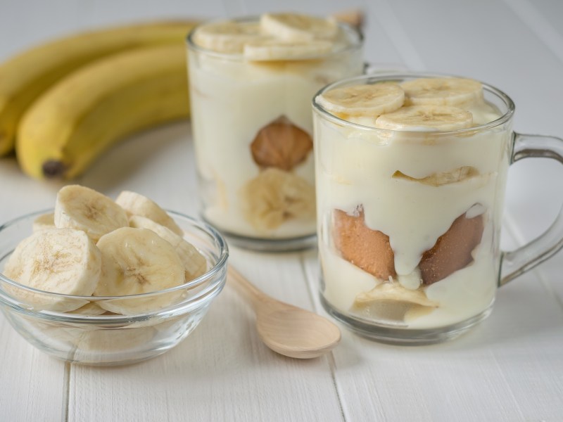 Zwei DessertglÃ¤ser mit Banana-Pudding. Daneben steht ein kleines SchÃ¤lchen mit Bananenscheiben. AuÃŸerdem liegen zwei Bananen und ein HolzlÃ¶ffel dabei.