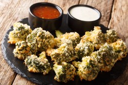 Ein schwarzer Teller mit Brokkoli-Bites aus dem Airfryer. Die Brokkoli-Bites sind mit zwei kleinen Schalen mit verschiedenen Dips serviert. Der Teller steht auf einer braunen Holzoberfläche.