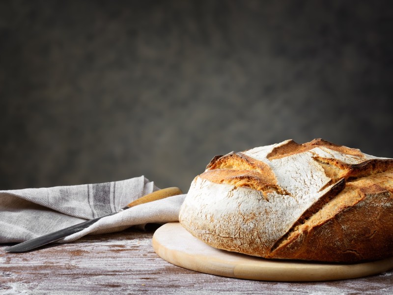 Ein Brot aus drei Zutaten liegt auf einem runden Brett auf einem schlichten Holztisch. Links daneben liegt ein Tuch sowie ein Brotmeser.