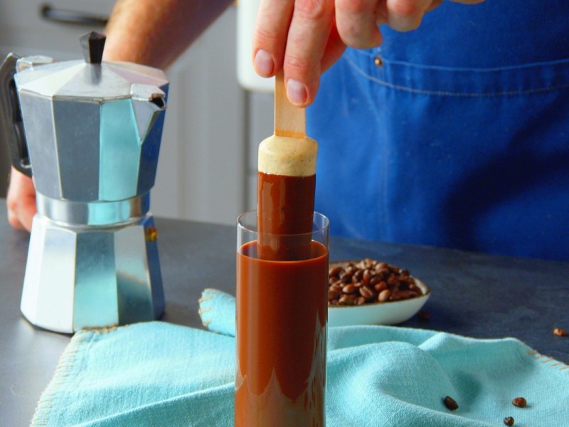 Ein Cheesecake am Stiel wird in ein Schnapsglas mit flÃ¼ssigher Schokolade getaucht.