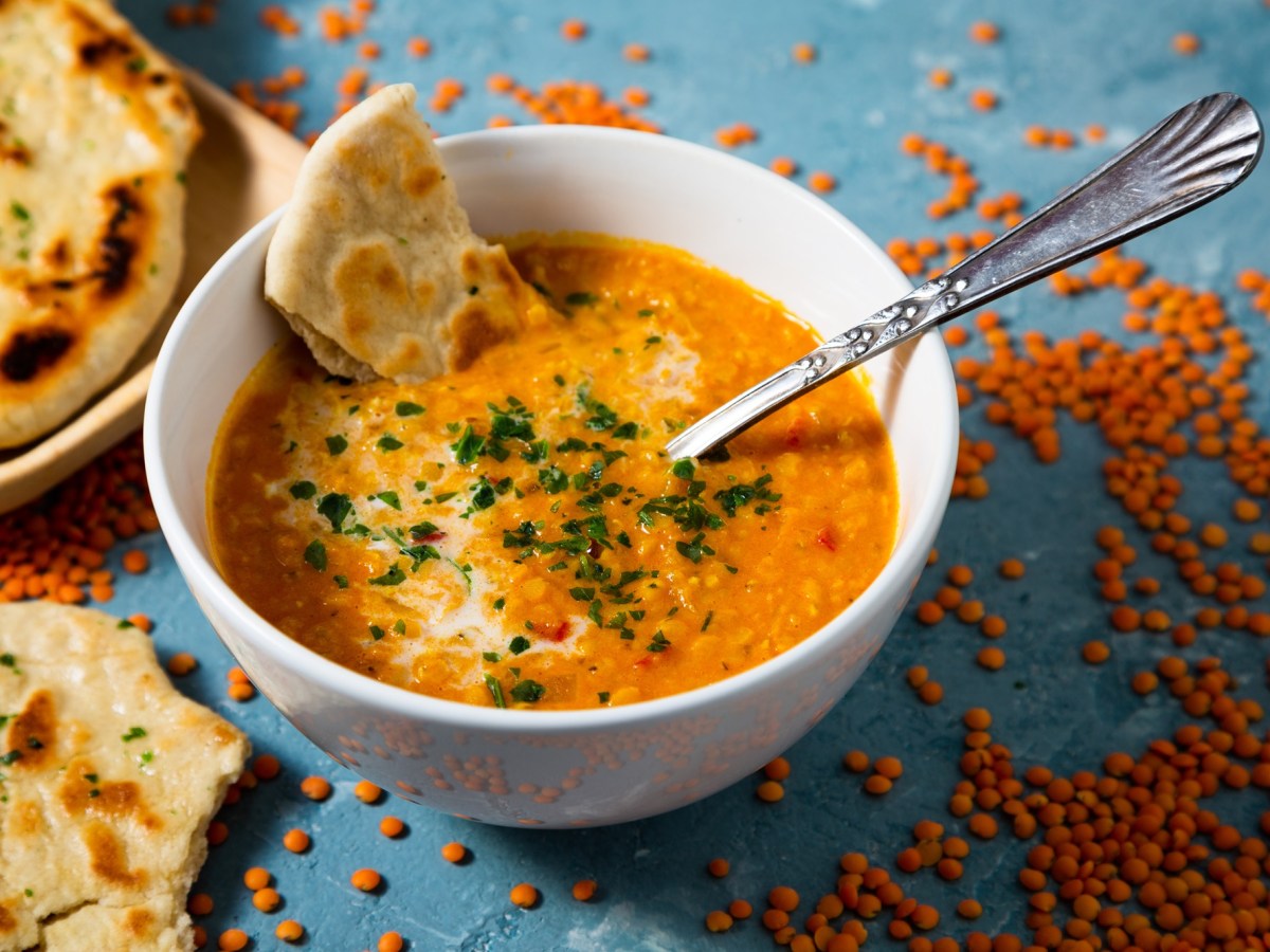 eine Schüssel Curry-Linsen-Suppe mit einem Stück Naanbrot und einem Löffel. Daneben rohe rote Linsen und Brot.