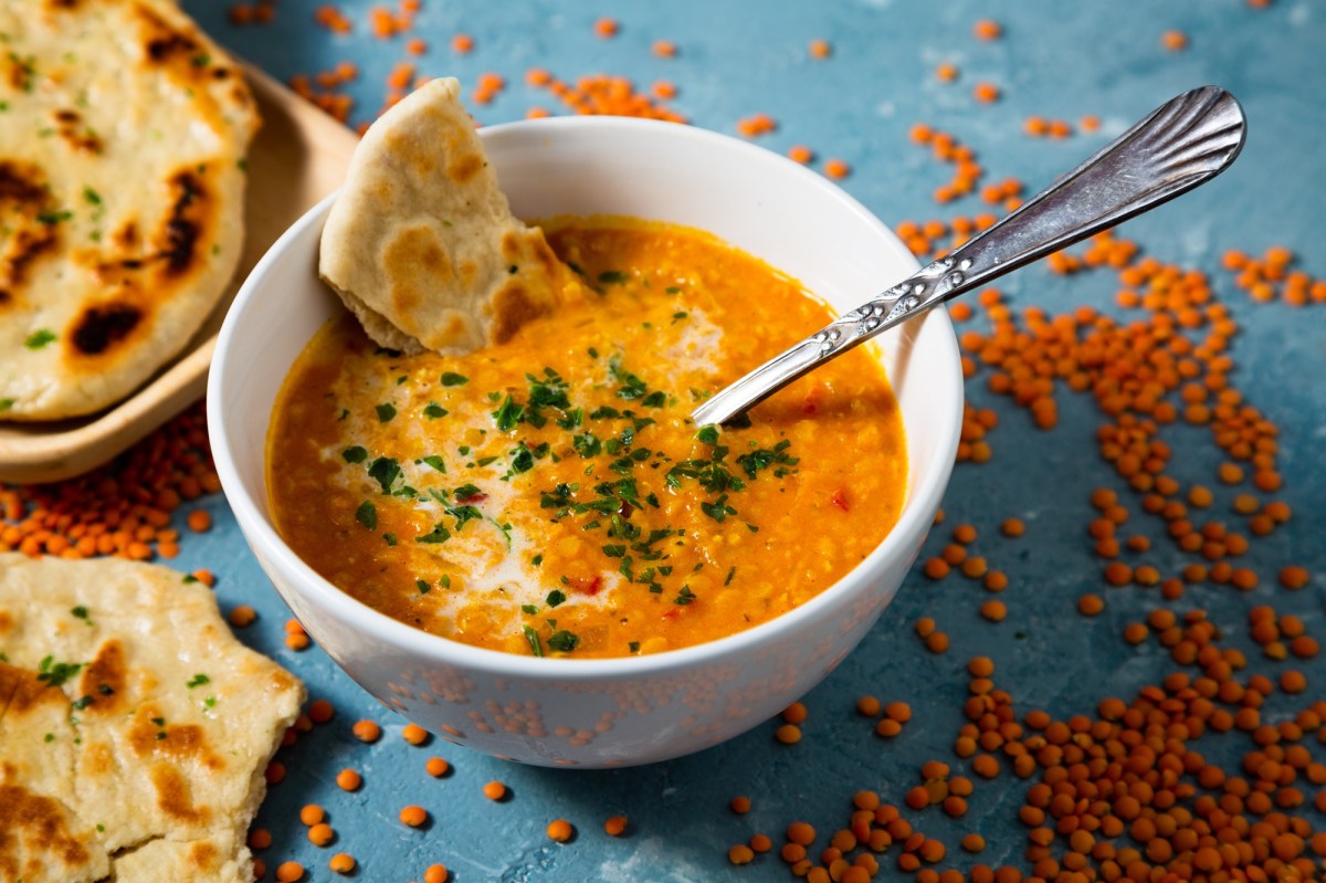 eine SchÃ¼ssel Curry-Linsen-Suppe mit einem StÃ¼ck Naanbrot und einem LÃ¶ffel. Daneben rohe rote Linsen und Brot.