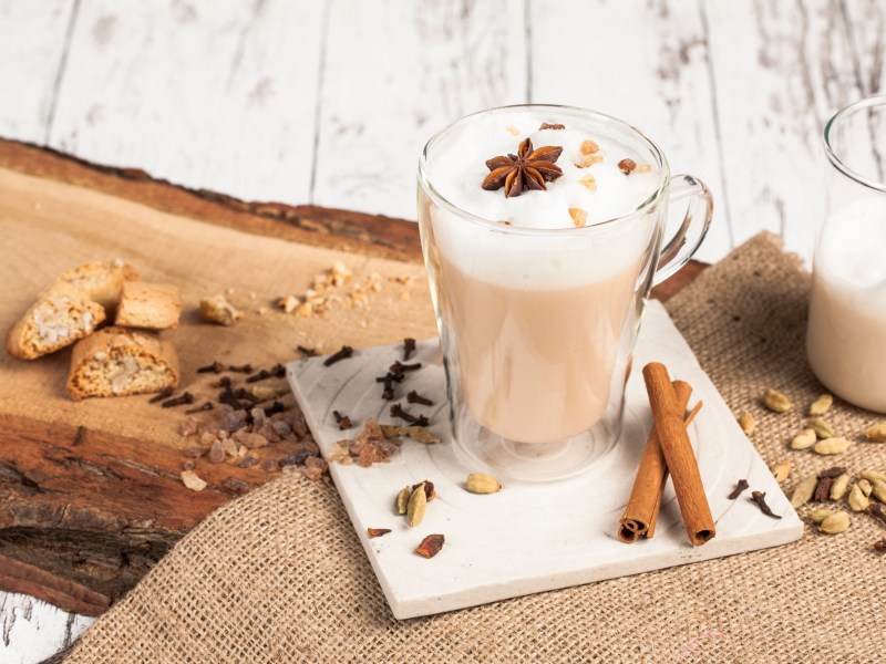 Eine Glastasse mit Dirty Chai Latte einem weiÃŸen Untersetzer. Der Untersetzer mit der Glastasse steht auf einem StÃ¼ck Holz, das mit Sackleinen bedeckt ist. AuÃŸerdem liegen Cantuccini rum, Zimtstangen, Nelken und andere Chai-GewÃ¼rze.