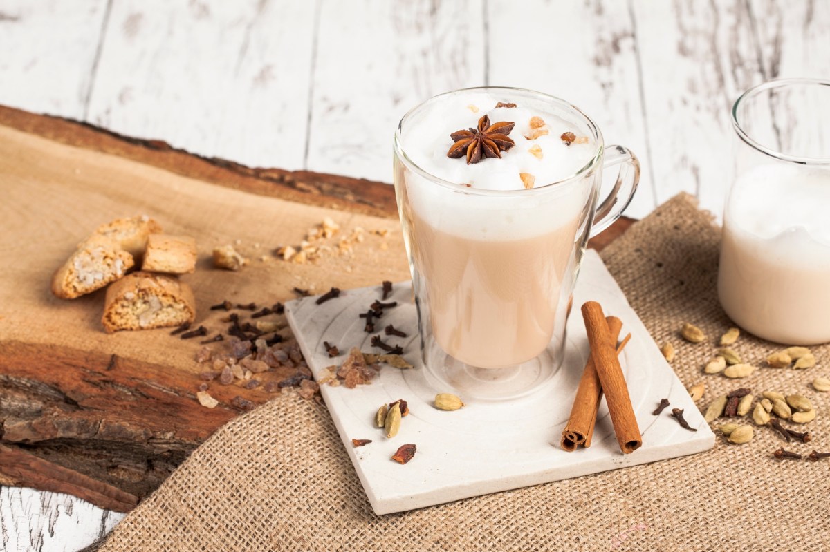 Eine Glastasse mit Dirty Chai Latte einem weiÃŸen Untersetzer. Der Untersetzer mit der Glastasse steht auf einem StÃ¼ck Holz, das mit Sackleinen bedeckt ist. AuÃŸerdem liegen Cantuccini rum, Zimtstangen, Nelken und andere Chai-GewÃ¼rze.