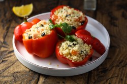 Drei gefüllte rote Paprikaschoten liegen auf einem weißen Teller. Der obere Teil ist als Deckel abgeschnitten. Sie sind mit Couscous, Oliven und Kräutern gefüllt. Im Hintergrund liegt eine Zitronenspalte.