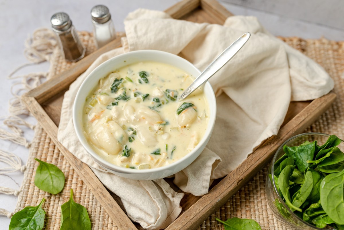 Eine SchÃ¼ssel voll cremiger Gnocchi-Suppe mit Artischocken und Spinat serviert auf einem Holzbrett ausgelegt mit einer beigen Serviette. Daneben ist ein Salz- und Pfefferstreuer zu sehen. AuÃŸerdem steht eine kleine Schale mit SpinatblÃ¤ttern daneben.