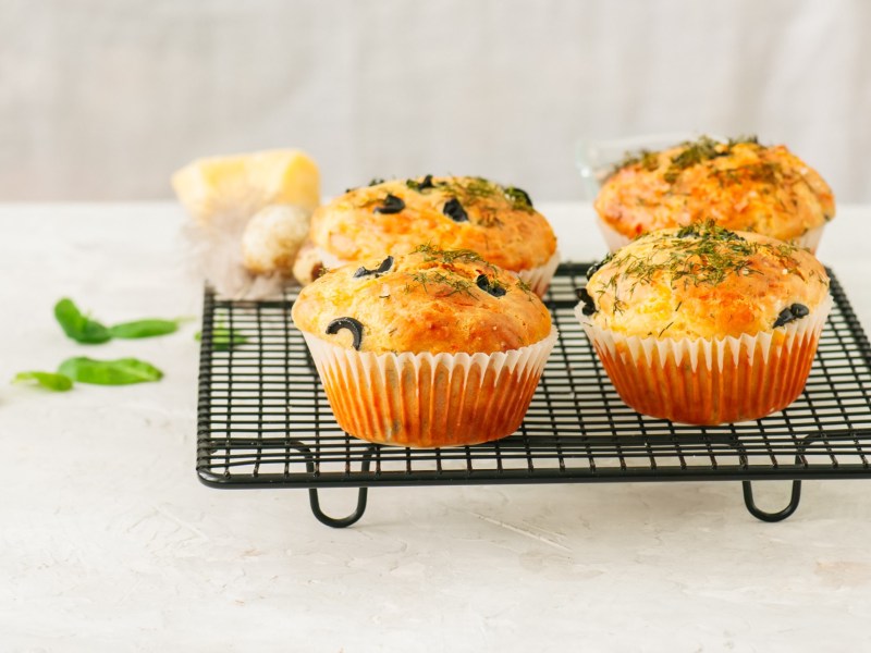 Herzhafte Haferfluckenmuffins mit KrÃ¤utern und schwarzen Olivenringen stehen auf einem schwarzen Gitter angerichtet. Im Hintergrund liegen ein paar frische BasilikumblÃ¤tter.