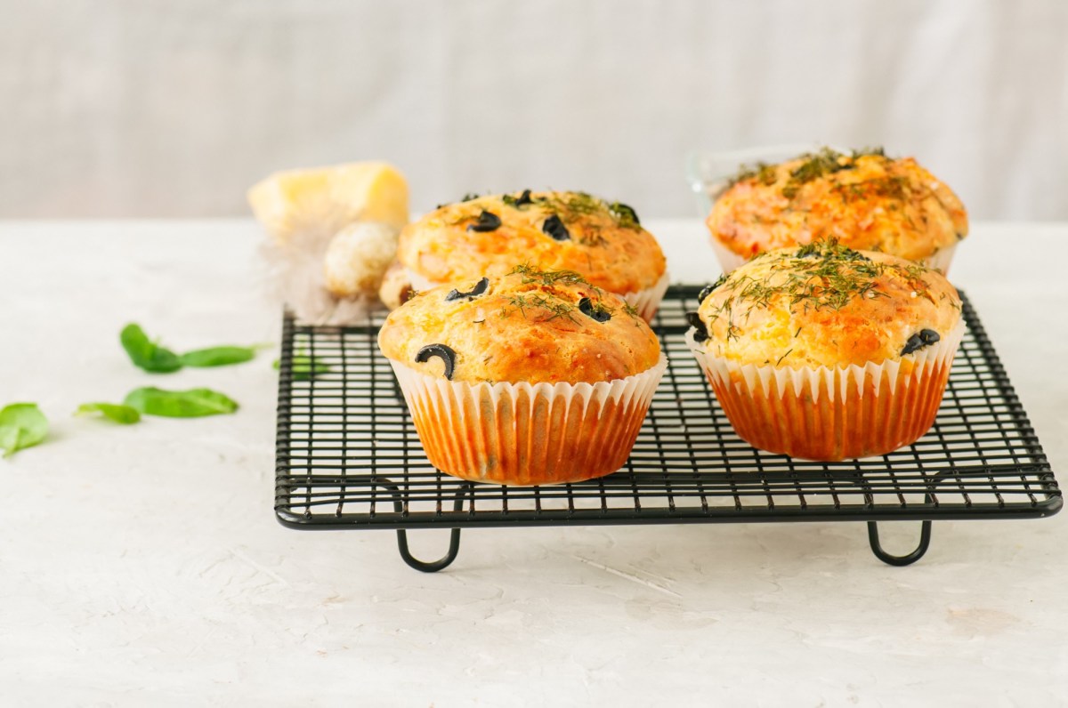 Herzhafte Haferfluckenmuffins mit KrÃ¤utern und schwarzen Olivenringen stehen auf einem schwarzen Gitter angerichtet. Im Hintergrund liegen ein paar frische BasilikumblÃ¤tter.