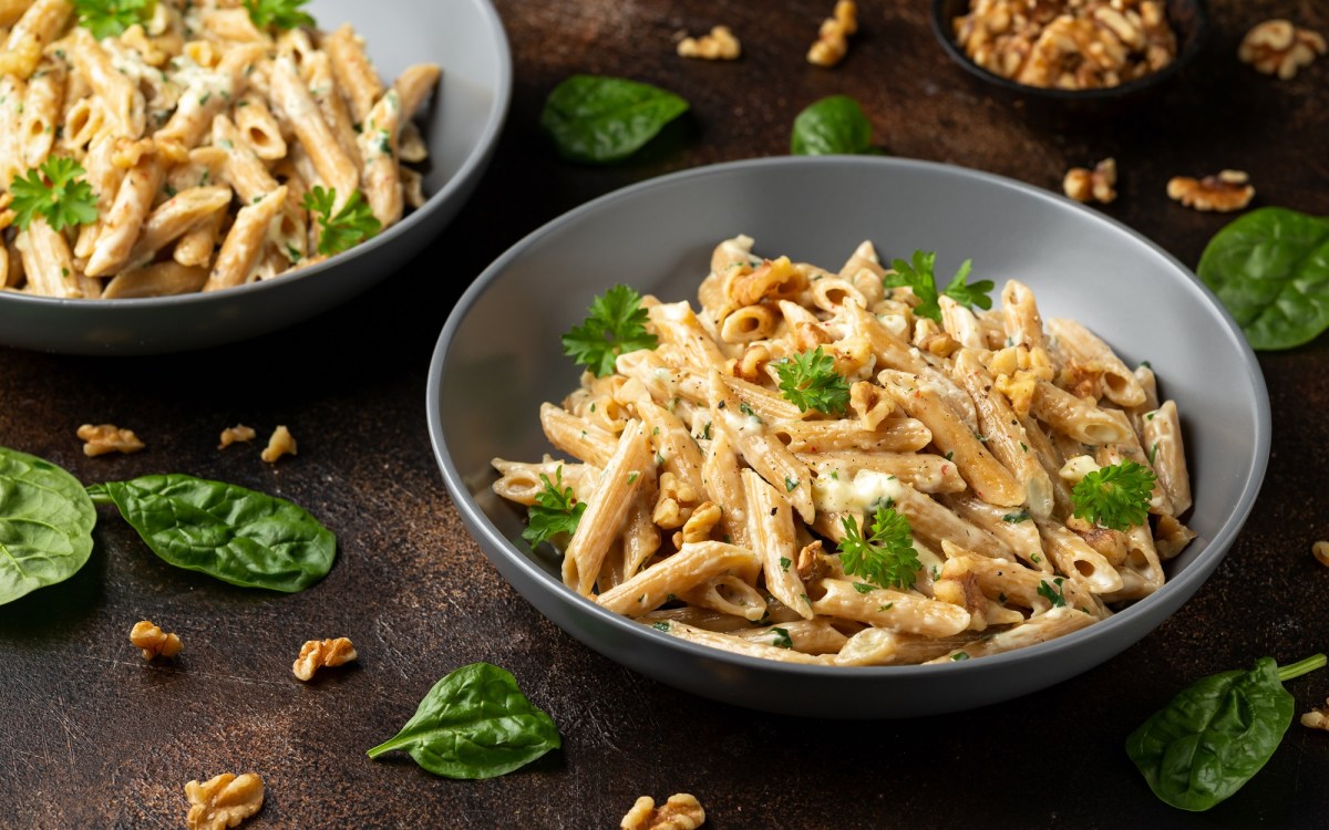 Ein grauer Teller mit einer groÃŸen Portion Hummus-Pasta, die mit frischer Petersilie und gehackten WalnÃ¼ssen serviert wird. Im Hintergrund ist eine weitere Portion des Gerichts zu sehen. Auf dem Tisch liegen BasilikumblÃ¤tter und WalnussstÃ¼ckchen verteilt.