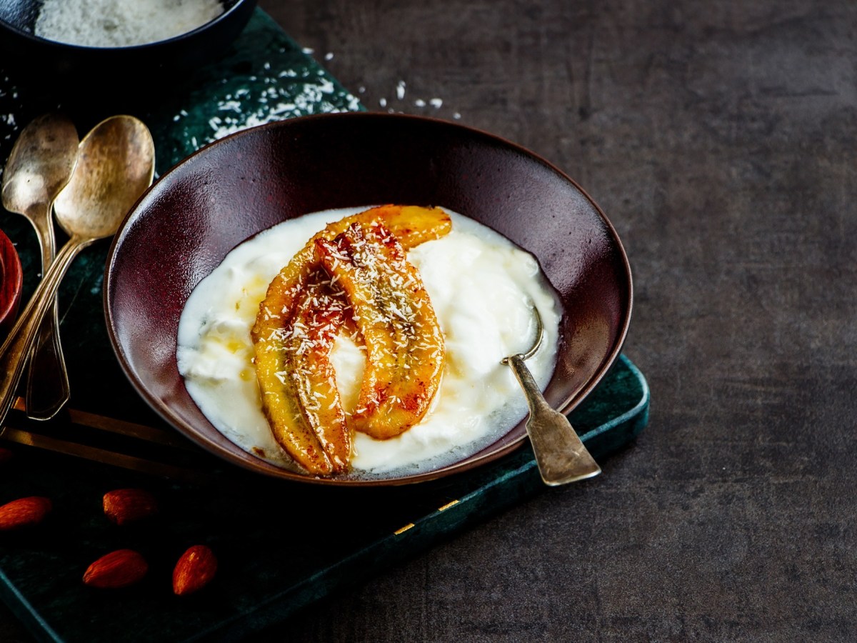 Eine Schüssel voll Joghurt mit karamellisierten Bananen. Garniert mit Kokosraspel. Daneben liegen Löffel und ein paar Gojibeeren.