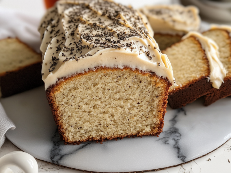 Ein London Fog Cake mit hellem ButterfrischkÃ¤se-Frosting und Earl Grey Tee bestreut. Der Kuchen ist kastenfÃ¶rmig und liegt angeschnitten auf einer Marmorplatte. Rechts daneben liegen ein paar weitere Scheiben London Fog Cake. Links auch eine Scheibe. AuÃŸerdem ist in der linken unteren Bildecke eine Tasse mit Earl Grey Teemischung zu sehen.