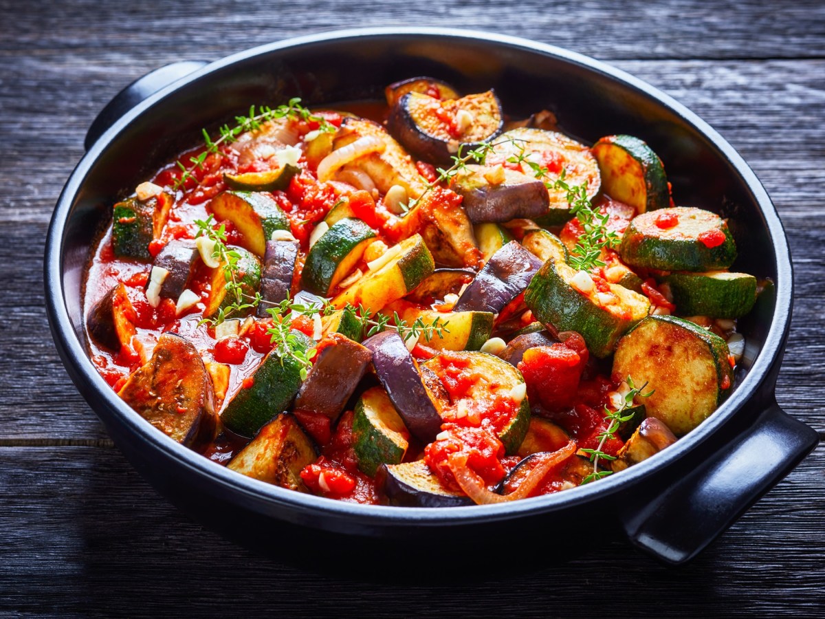 Ein Gemüseeintopf mit Auberginen, Zucchini, Tomaten, Knoblauch und Zwiebeln, serviert in einer schwarzen Keramikschale und garniert mit Kräutern. Die Schale steht auf einem dunklen Holzuntergrund