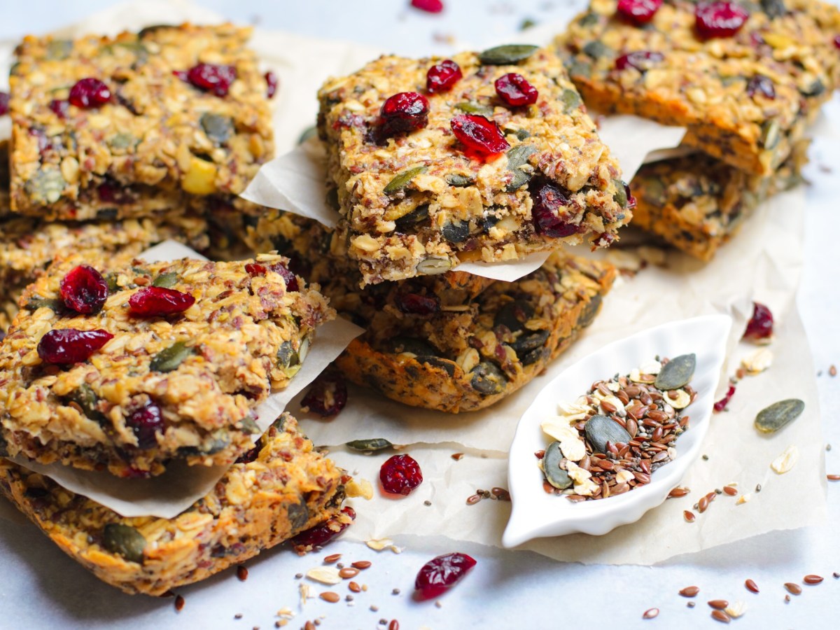 Einige MÃ¼sliriegel mit Cranberrys, KÃ¼rbiskernen und Kokos, die auf einer weiÃŸen ArbeitsflÃ¤che in kleinen Stapeln angerichtet sind. Es liegen getrocknete Cranberrys, KÃ¼rbiskerne und Leinsamen verstreut.