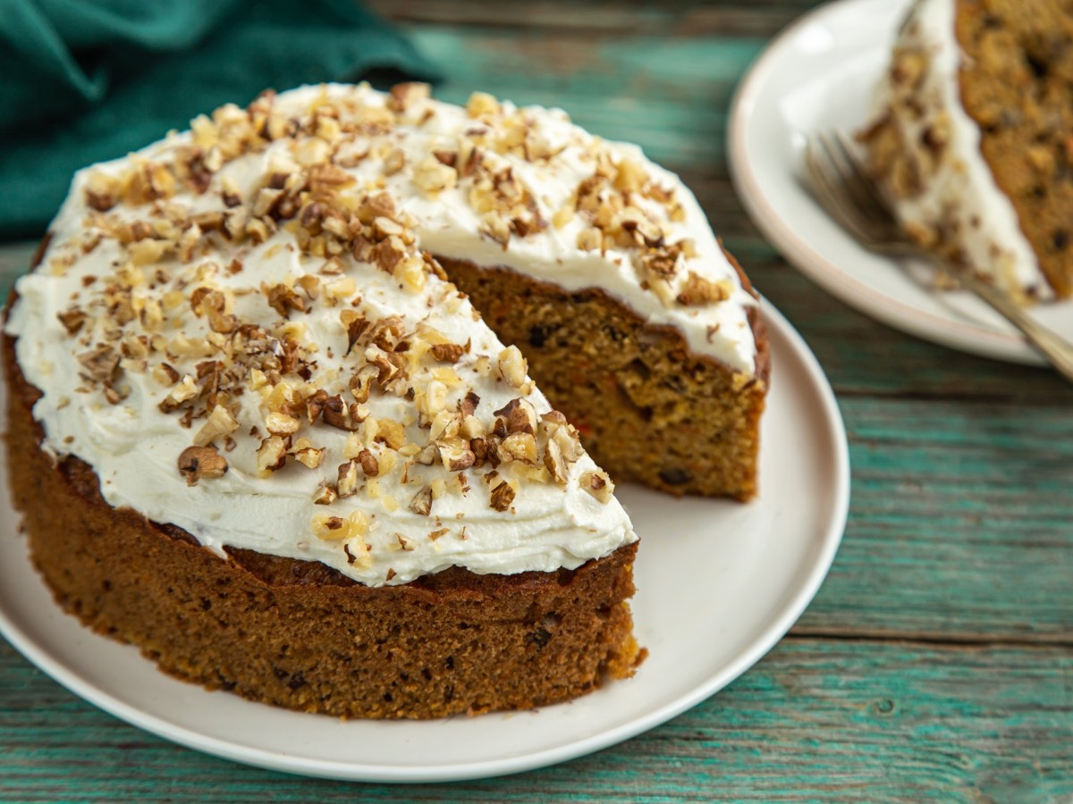 Ein runder Nusskuchen ohne Mehl steht auf einem weiÃŸen Teller auf einem grÃ¼nlichen Holztisch. Er ist mit weiÃŸem Frosting versehen und mit NussstÃ¼ckchen dekoriert. Im Hintergrund steht ein StÃ¼ck des Kuchens.