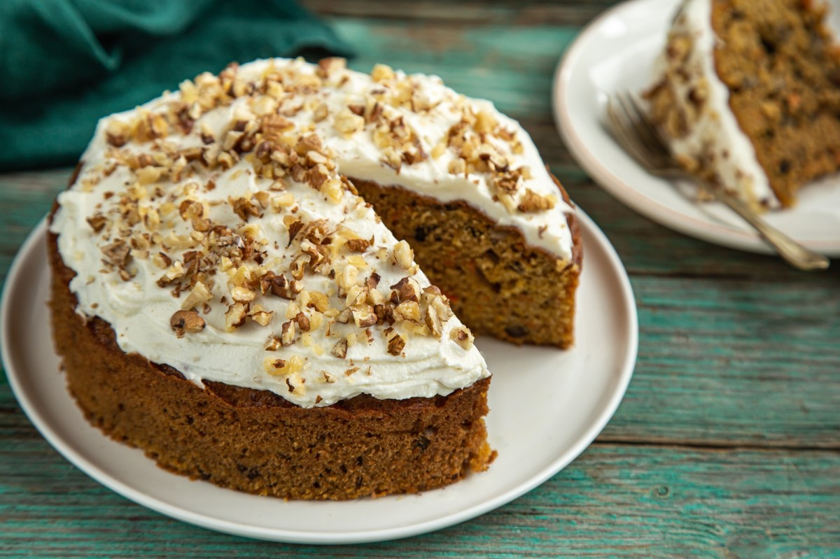 Ein runder Nusskuchen ohne Mehl steht auf einem weiÃŸen Teller auf einem grÃ¼nlichen Holztisch. Er ist mit weiÃŸem Frosting versehen und mit NussstÃ¼ckchen dekoriert. Im Hintergrund steht ein StÃ¼ck des Kuchens.