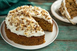 Ein runder Nusskuchen ohne Mehl steht auf einem weißen Teller auf einem grünlichen Holztisch. Er ist mit weißem Frosting versehen und mit Nussstückchen dekoriert. Im Hintergrund steht ein Stück des Kuchens.