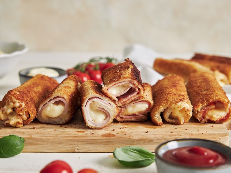 Sieben panierte ToastrÃ¶llchen, gefÃ¼llt mit Schhinken und KÃ¤se liegen auf einem Holzbrett. Im Hintergeund liegen weitere RÃ¶llchen, Tomaten und Mayonnaise. Im Vordergrund sieht man ein paar BasilikumblÃ¤tter, eine Schale mit Ketchup und zwei Tomaten.