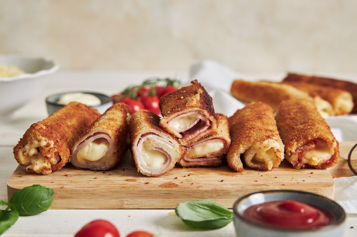 Sieben panierte ToastrÃ¶llchen, gefÃ¼llt mit Schhinken und KÃ¤se liegen auf einem Holzbrett. Im Hintergeund liegen weitere RÃ¶llchen, Tomaten und Mayonnaise. Im Vordergrund sieht man ein paar BasilikumblÃ¤tter, eine Schale mit Ketchup und zwei Tomaten.