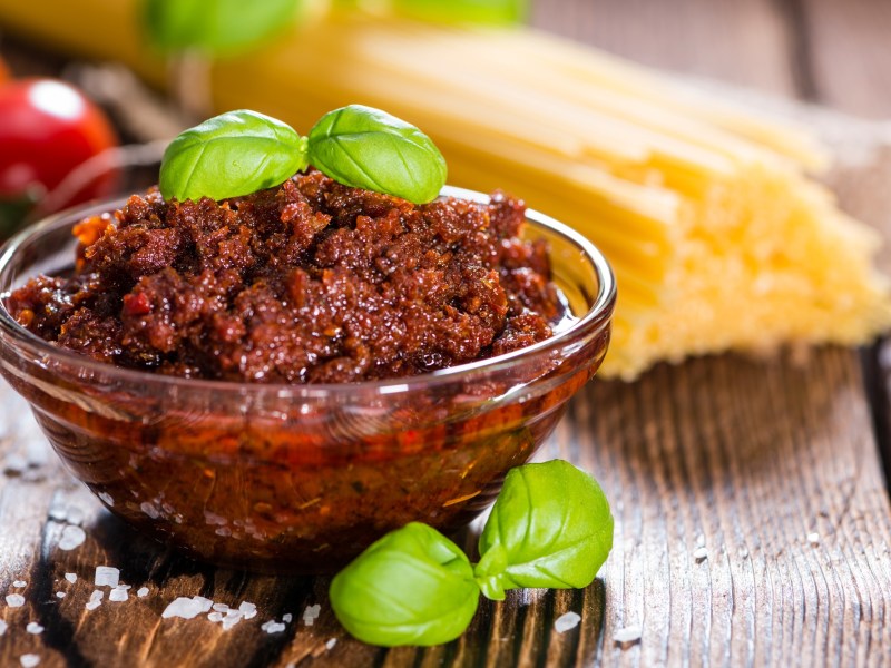 Eine Glasschale Pesto Rosso garniert mit Basilikum, dahinter unscharf Tomaten und rohe Spaghetti.