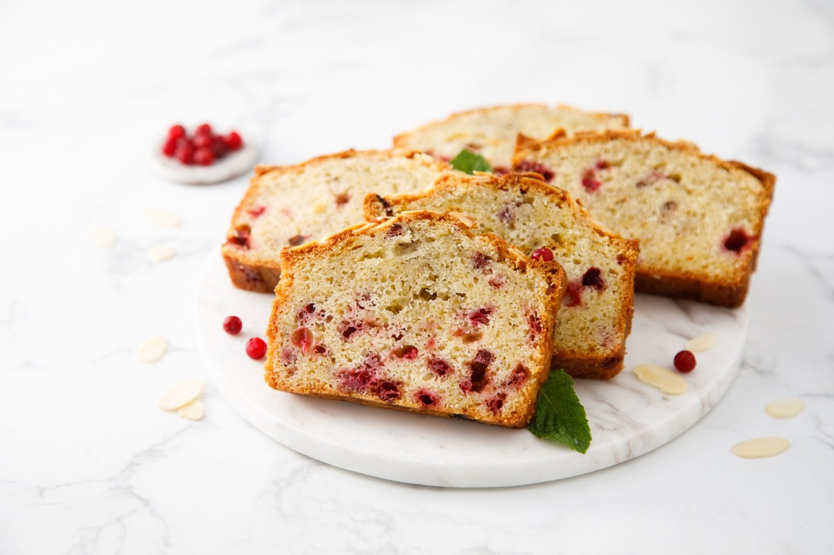 Eine heller marmorner Teller mit fÃ¼nf Scheiben Preiselbeerkuchen. Als Garnitur liegen frische MinzblÃ¤tter dabei. Ringsum liegen einige Mandelsplitter und frische Preisebeeren. Im Hintergrund ist ein kleines SchÃ¤lchen mit Preiselbeeren zu sehen.