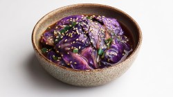 Eine hellbraune Keramikschüssel mit Rotkohl-Kimchi, das mit Sesam bestreut ist. Die Schüssel steht auf einer hellen Arbeitsfläche.