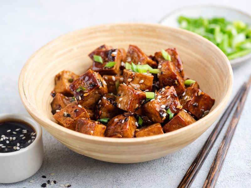 Teriyaki Tofu aus dem Airfryer in WÃ¼rfel geschnitten in einer kleinen Bambusschale. Sie sind mit FrÃ¼hlingszwiebeln und hellem sowie schwarzem Sesam angerichtet. Im Hintergrund steht ein kleiner Teller mit geschnittenen FrÃ¼hlingszwiebeln. Im Vordergrund steht eine Schale mit SojasoÃŸe.