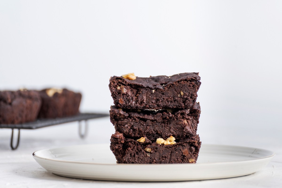 Drei StÃ¼cke vegane Kichererbsen-Brownies auf einem weiÃŸen groÃŸen Teller aufeinander gestapelt. Auf einem Gitter im Hintergrund sind weitere vegane Kichererbsen-BrowniestÃ¼cke zu sehen.