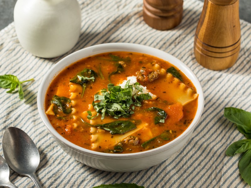 Lasagnesuppe in einer weiÃŸen SchÃ¼ssel steht auf einer gesteiften Tischdecke. Drumherum liegen ein LÃ¶ffel, BasilikumblÃ¤tter sowie ein Pfeffer- und Salzstreuer. Die Suppe ist ebenfalls mit Basilikum und KÃ¤se garniert.