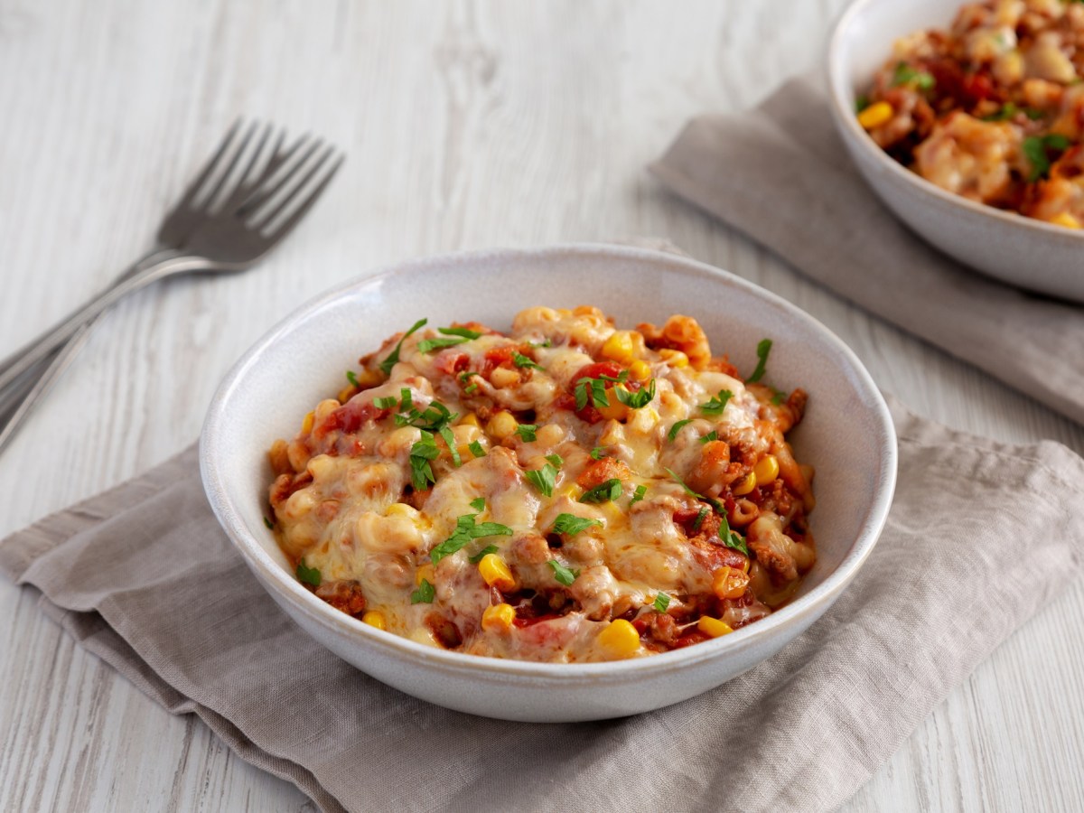 Ein weißer Teller voll mit veganer One Pot Taco Pasta, der auf einer grauen Stoffserviette steht. Schräg links daneben liegen zwei Gabeln. In der rechten oberen Bildecke kann mit verschwommen einen weiteren Teller mit veganer One Pot Taco Pasta erkennen.