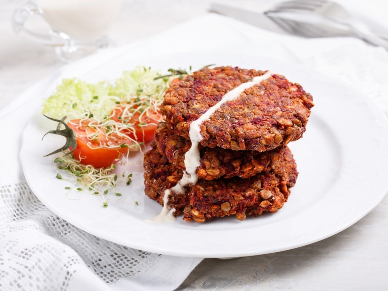 Vegane Rotkohlbratlinge mit Haferflocken liegen auf einem weiÃŸen Teller. Daneben liegt etwas Gartenkresse und eine halbierte Tomate.