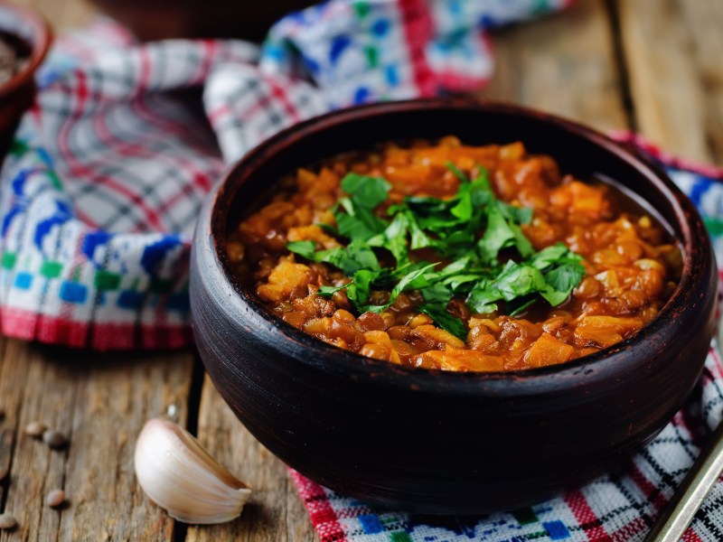 Eine SchÃ¼ssel veganer Linseneintopf mit KrÃ¤utern garniert, drumherum Knoblauch und ein Geschirrtuch.