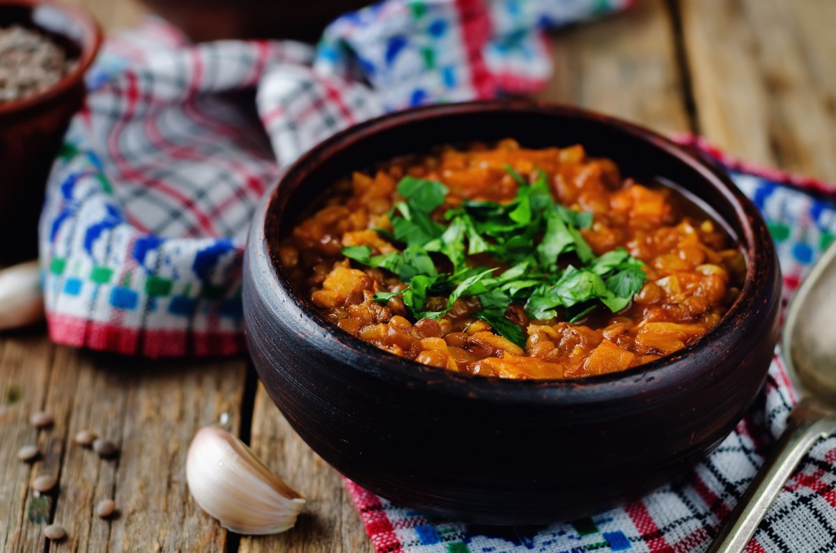 Eine SchÃ¼ssel veganer Linseneintopf mit KrÃ¤utern garniert, drumherum Knoblauch und ein Geschirrtuch.