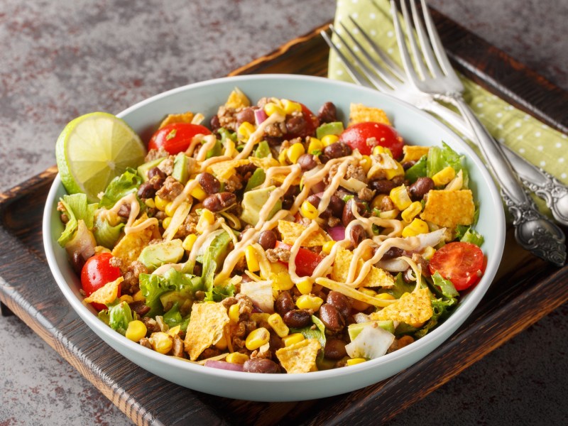 Bunter und veganer Nacho-Salat in einer weiÃŸen SchÃ¼ssel auf einem dunklen Holzbrett. Der Salat besteht aus Nachos, Kidneybohnen, Nachos, frischem Salat, Mais und Tomaten. Links neben der Schale liegen zwei Gabeln auf einer Serviette.