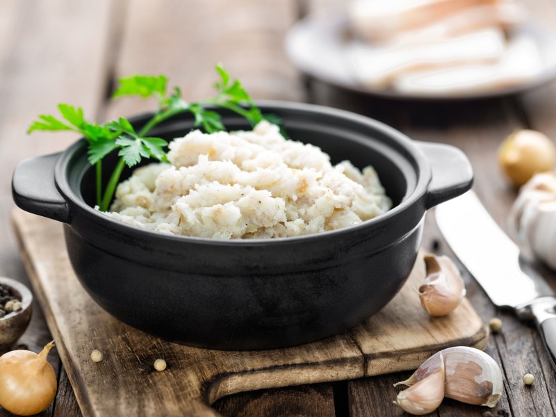 Ein schwarzes TÃ¶pfchen mit veganem Schmalz, garniert mit frischer Petersilie, angerichtet auf einem Holzbrett. Daneben liegt ein Messer, sowie Knoblauchzehen und PfefferkÃ¶rner.