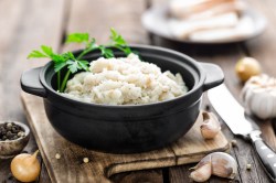 Ein schwarzes Töpfchen mit veganem Schmalz, garniert mit frischer Petersilie, angerichtet auf einem Holzbrett. Daneben liegt ein Messer, sowie Knoblauchzehen und Pfefferkörner.