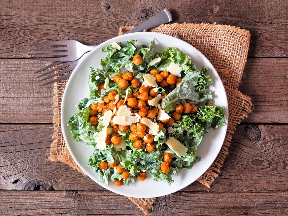 Draufsicht auf einen weißen Teller mit einer Portion vegetarischem Ceasar Salad mit gerösteten Kichererbsen und Parmesanflocken. Der Teller steht auf einem Holztisch auf einer hellbraunen rustikalen Stoffserviette. Daneben liegen zwei Gabeln.