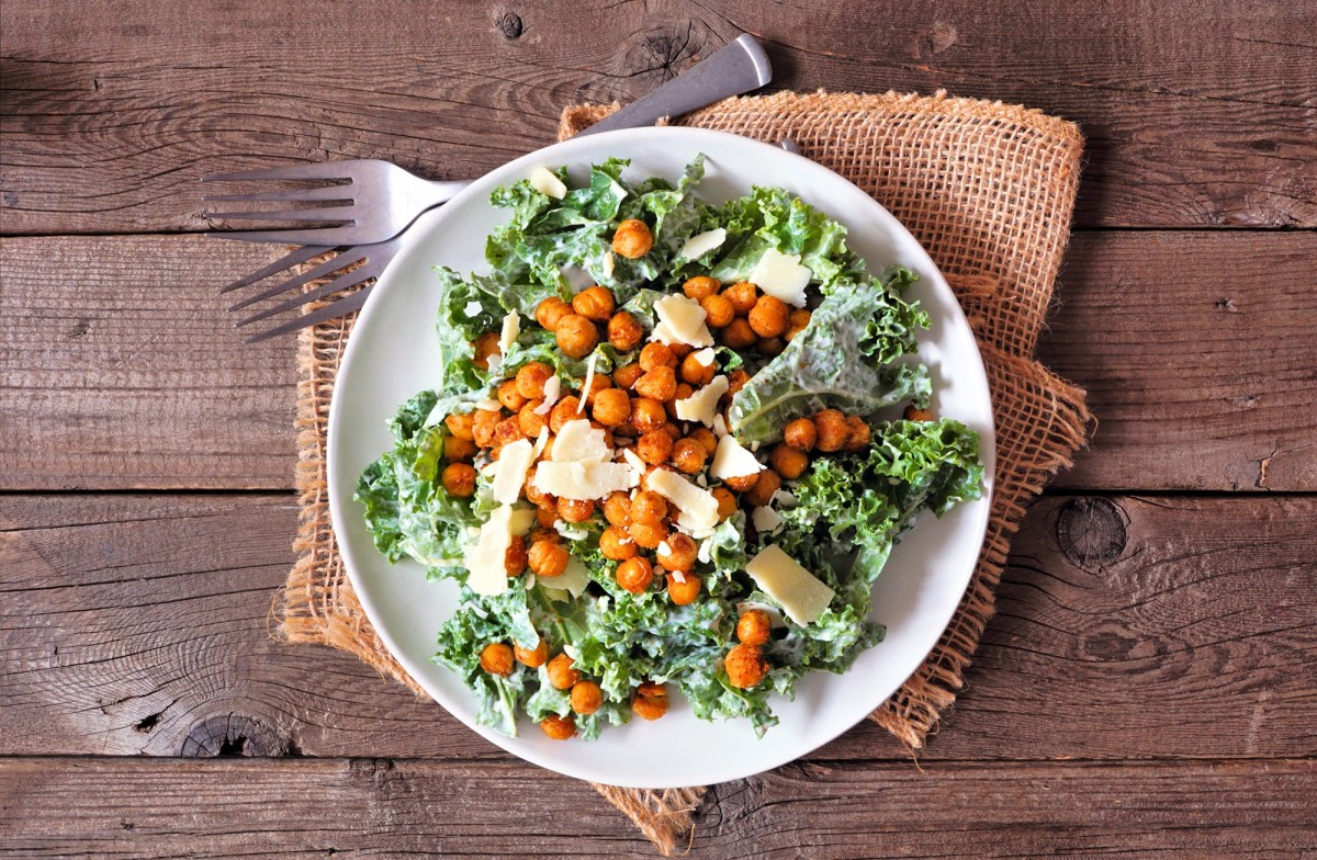 Draufsicht auf einen weiÃŸen Teller mit einer Portion vegetarischem Ceasar Salad mit gerÃ¶steten Kichererbsen und Parmesanflocken. Der Teller steht auf einem Holztisch auf einer hellbraunen rustikalen Stoffserviette. Daneben liegen zwei Gabeln.