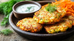 Fünf Zucchini-Dill-Bratlinge liegen auf einem dunklen Teller. Sie sind mit frischem Dill dekoriert. Auf dem Teller steht außerdem eine Schale mit Joghurt-Dill-Dip, im Hintergrund liegen geraspelte Möhrenstreifen.