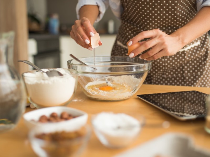 Tipps Backen: Eine Frau schlägt Eier in eine Schüssel auf. Drumherum stehen Backzutaten.