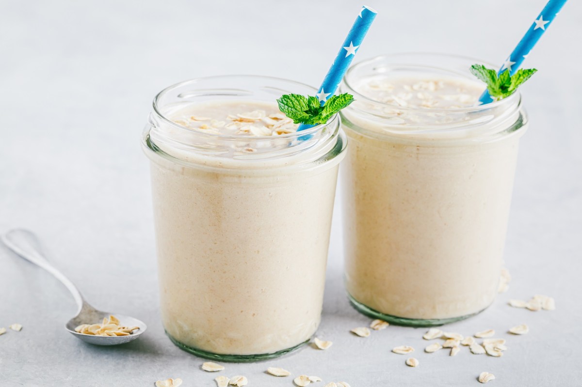 Zwei Gläser Käsekuchen-Shake mit blauen Strohhalmen und frischer Minze dekoriert. Die Käsekuchen-Shakes sind jeweils mit ein paar Haferflocken bestreut. Neben den Gläsern liegt ein Löffel mit Haferflocken und ringsum sind auch welche verstreut. Die Gläser stehen auf einer weißen Holzoberfläche.