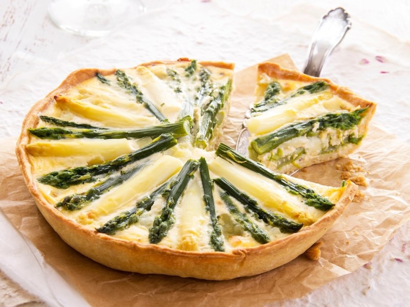 Eine runde, angeschnittene vegane Spargelquiche, aus der ein Stück herausgenommen wird, auf einem Stück Backpapier.