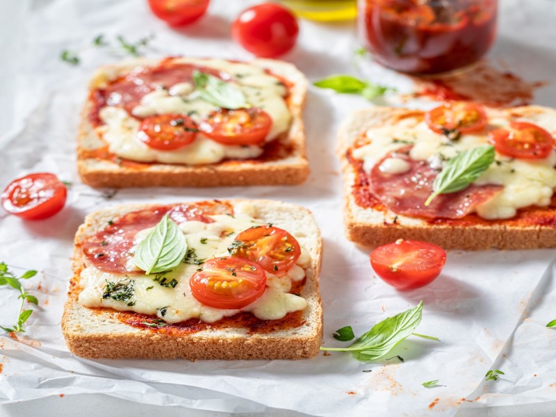 3 Pizza-Toasts aus dem Airfryer auf einem Stück Backpapier, belegt mit Käse, Tomaten und Salami.